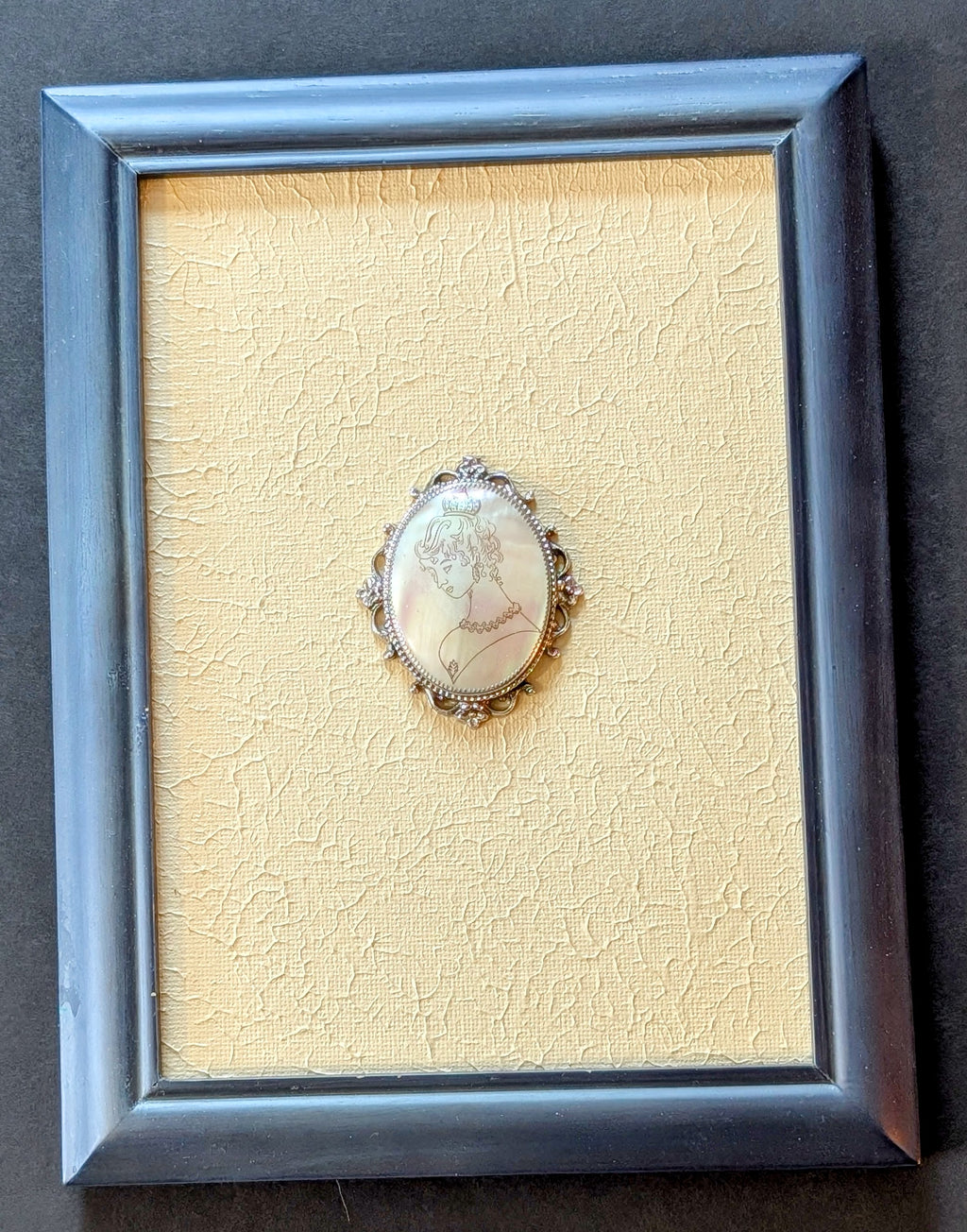 Framed jewelry piece with a detailed brooch on a textured beige background