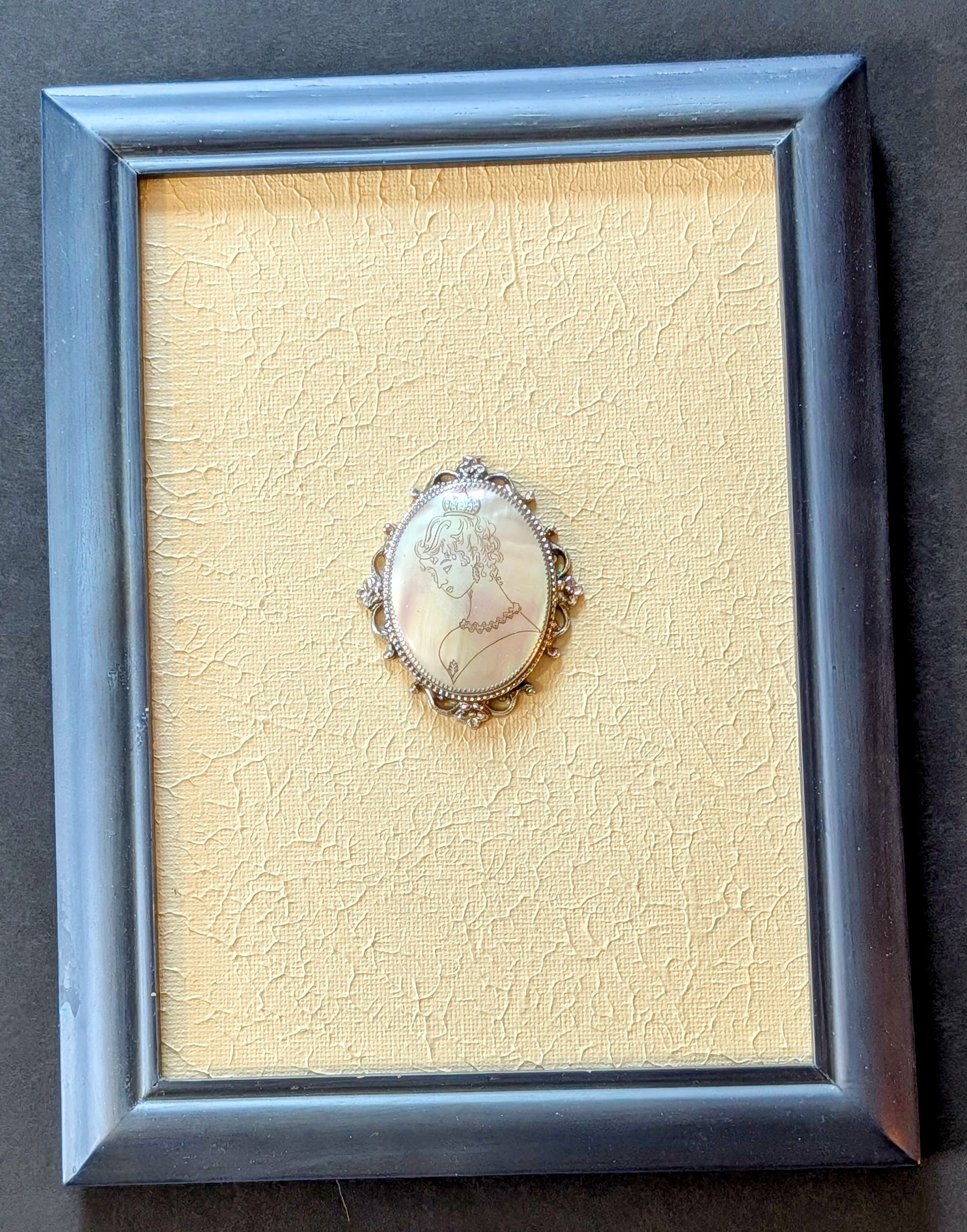 Framed jewelry piece with a detailed brooch on a textured beige background