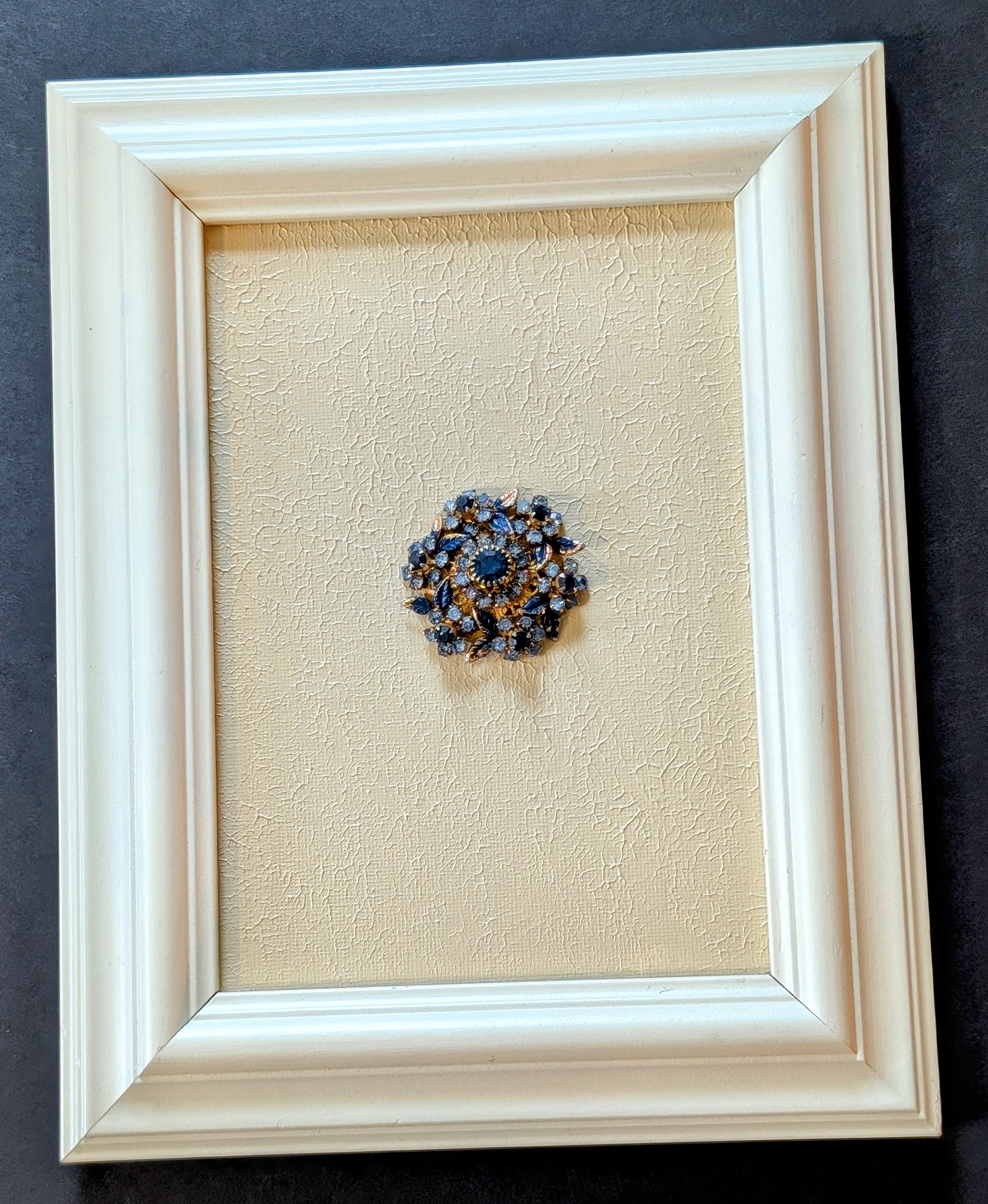 Framed decorative brooch with blue and gold elements on a textured beige background