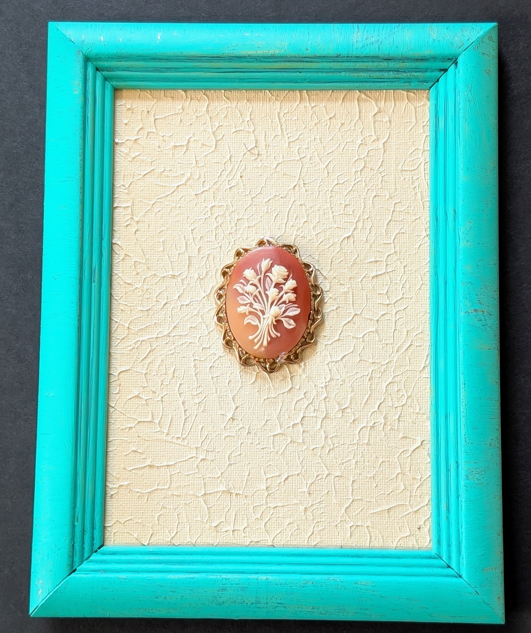 Coral-colored brooch with white design in a turquoise frame on a textured beige background