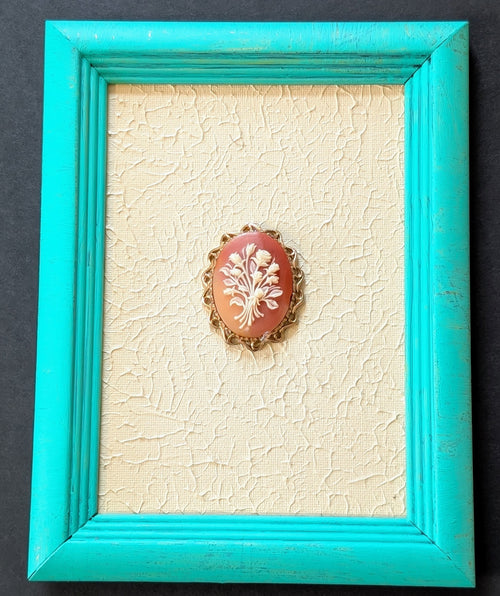 Coral-colored brooch with white design in a turquoise frame on a textured beige background
